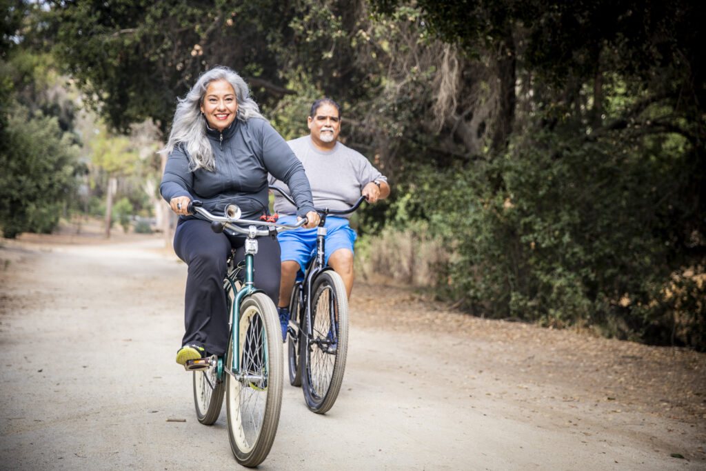 Older Hispanic couple riding bikes down a path surrounded by woods.
