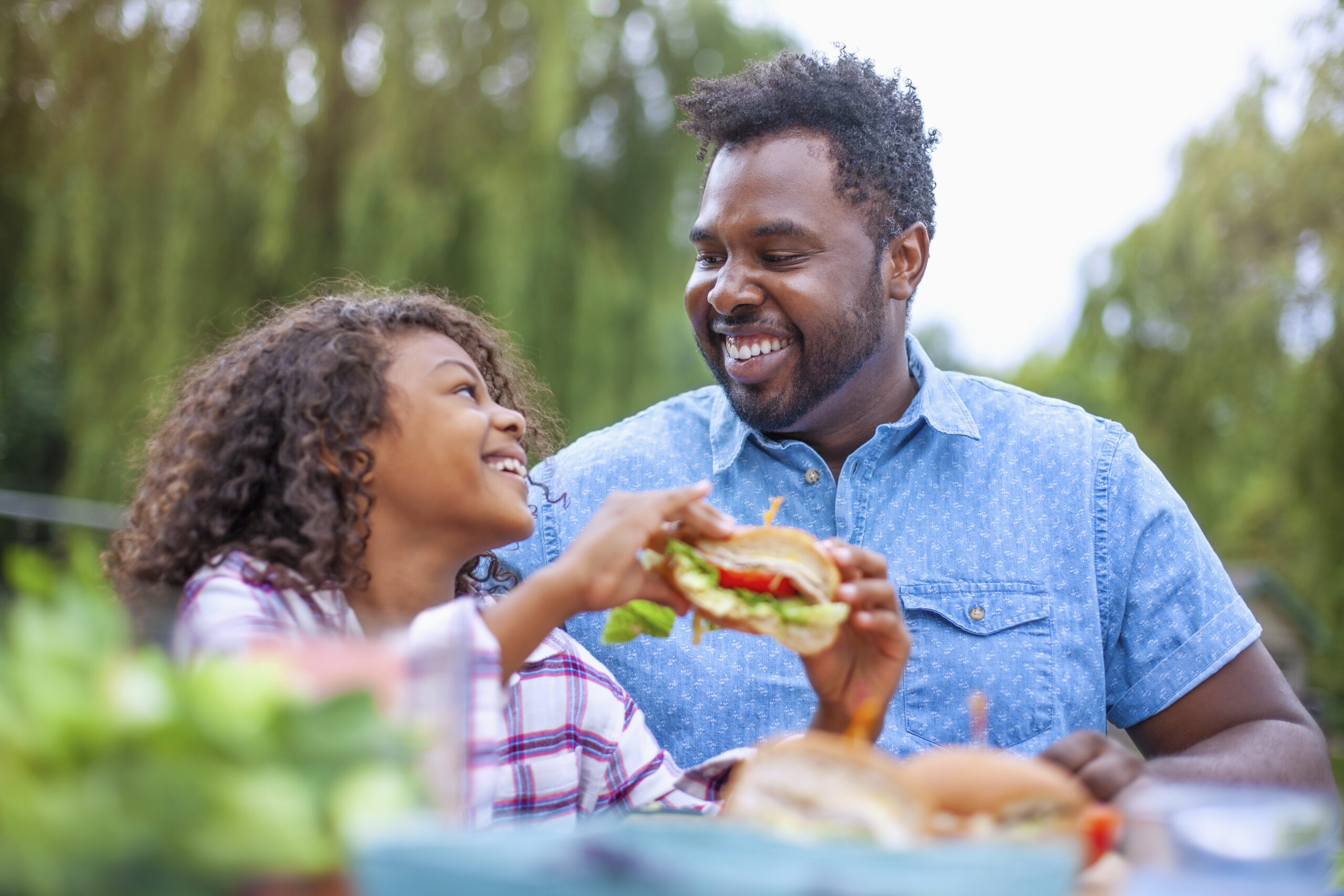 Dad and young daughter smiling and eating a burger outside at a picnic.
